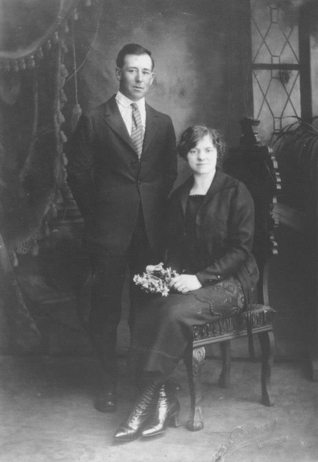Wedding photo of Robert and Anna. Robert is standing and wears a dark suit, white shirt and striped tie. Anna has short hair and is seated, wearing dark outfit with a long narrow skirt, long jacket and calf length laced boots.